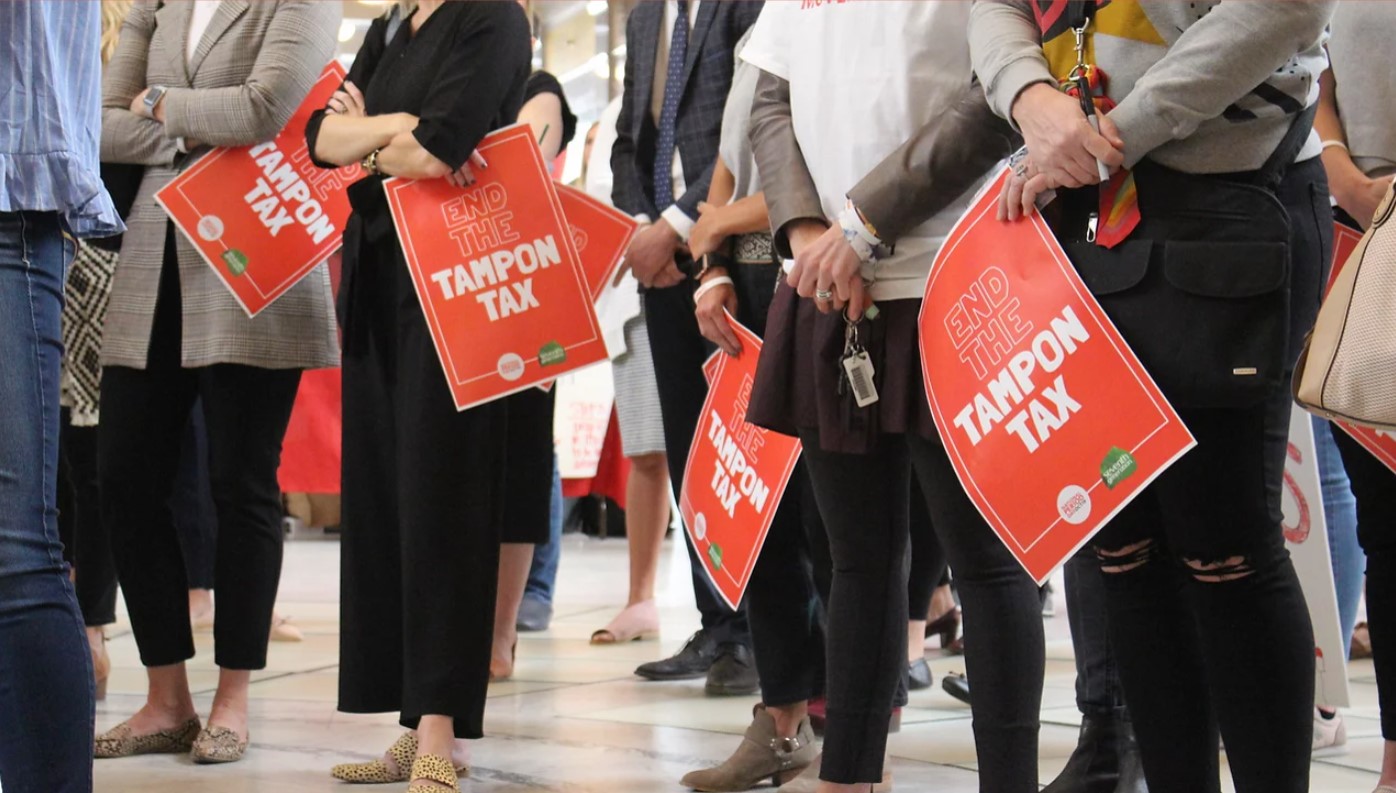 group of females shown from the arms down holding red posters that read 'Tampon Tax'