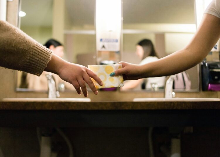 a hand passing a pad to another hand in a restroom with the girls' reflections out of focus in the mirror